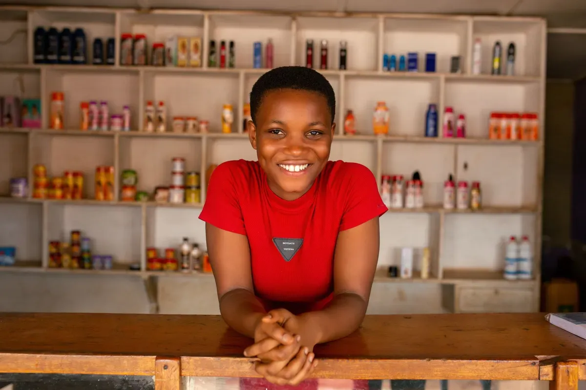 Dona de uma pequena loja sorrindo atrás do balcão, com as prateleiras abastecidas ao fundo.