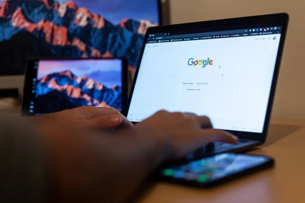 Pessoa digitando no notebook aberto na página do Google, com o celular ao lado